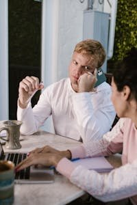 A businessman in a white hoodie looks puzzled on a call while a business analyst types on a laptop outdoors.