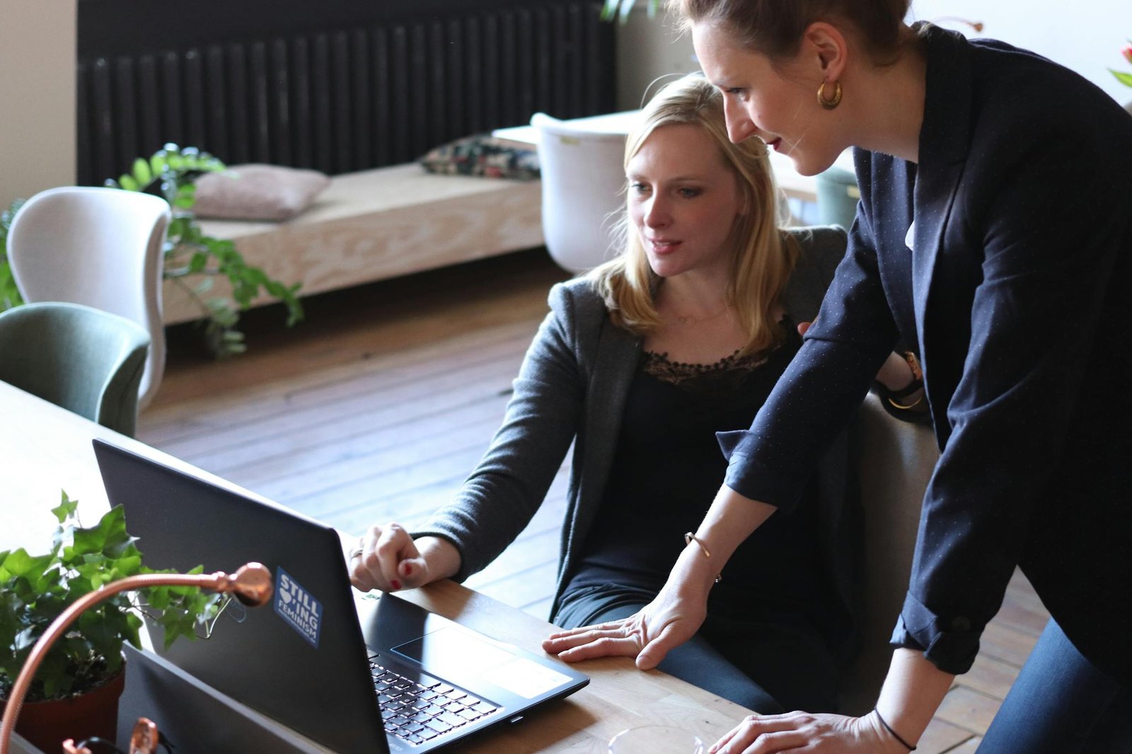 Two professional women discuss a website project using a laptop in a modern office environment.