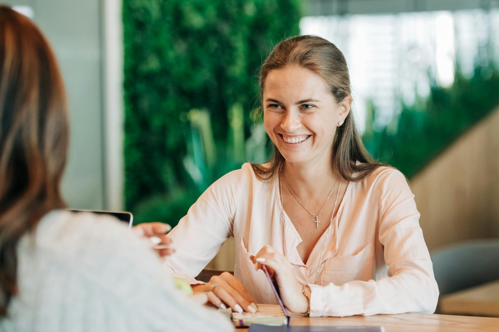 A young local Irish businesswoman surrounding with green background of plants smiling facing another businesswoman on her laptop reviewing a website